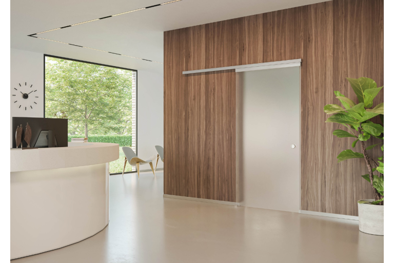 Een gesloten glazen deur in een moderne receptieruimte, die leidt naar een ruimte met twee stoelen. De deur bevindt zich in een houten wandpaneel achter een gebogen witte balie.